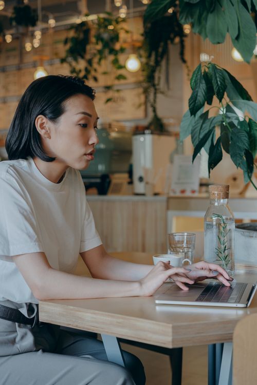 Free Asian woman typing on a laptop in a coffee shop, embodying the digital nomad lifestyle. Stock Photo