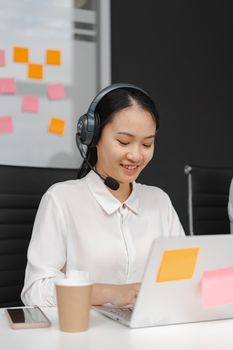 Smiling woman in a call center environment with headphones, working on a laptop indoors.