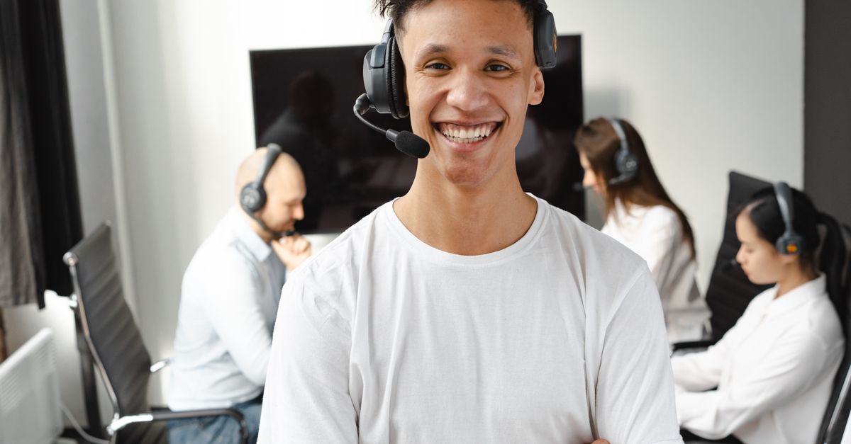 Young call center agent smiling confidently while colleagues work in the background, showcasing teamwork and customer service.