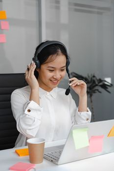 Smiling Asian woman in white shirt working as a call center agent with laptop and headset in office.