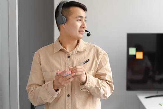 Asian man in a call center setting wearing a headset, holding notes and a pen indoors.