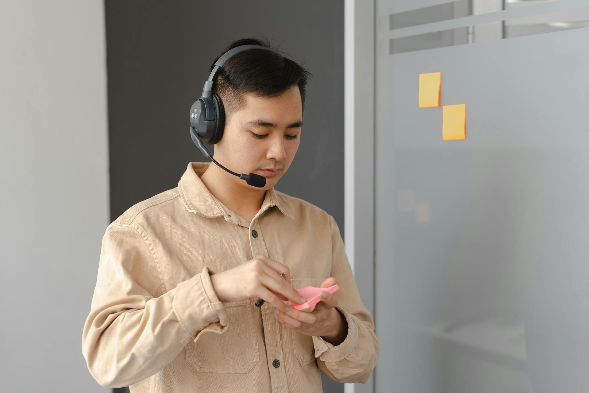 Young adult in an office setting using a headset and sticky notes. Ideal for business communication visuals.