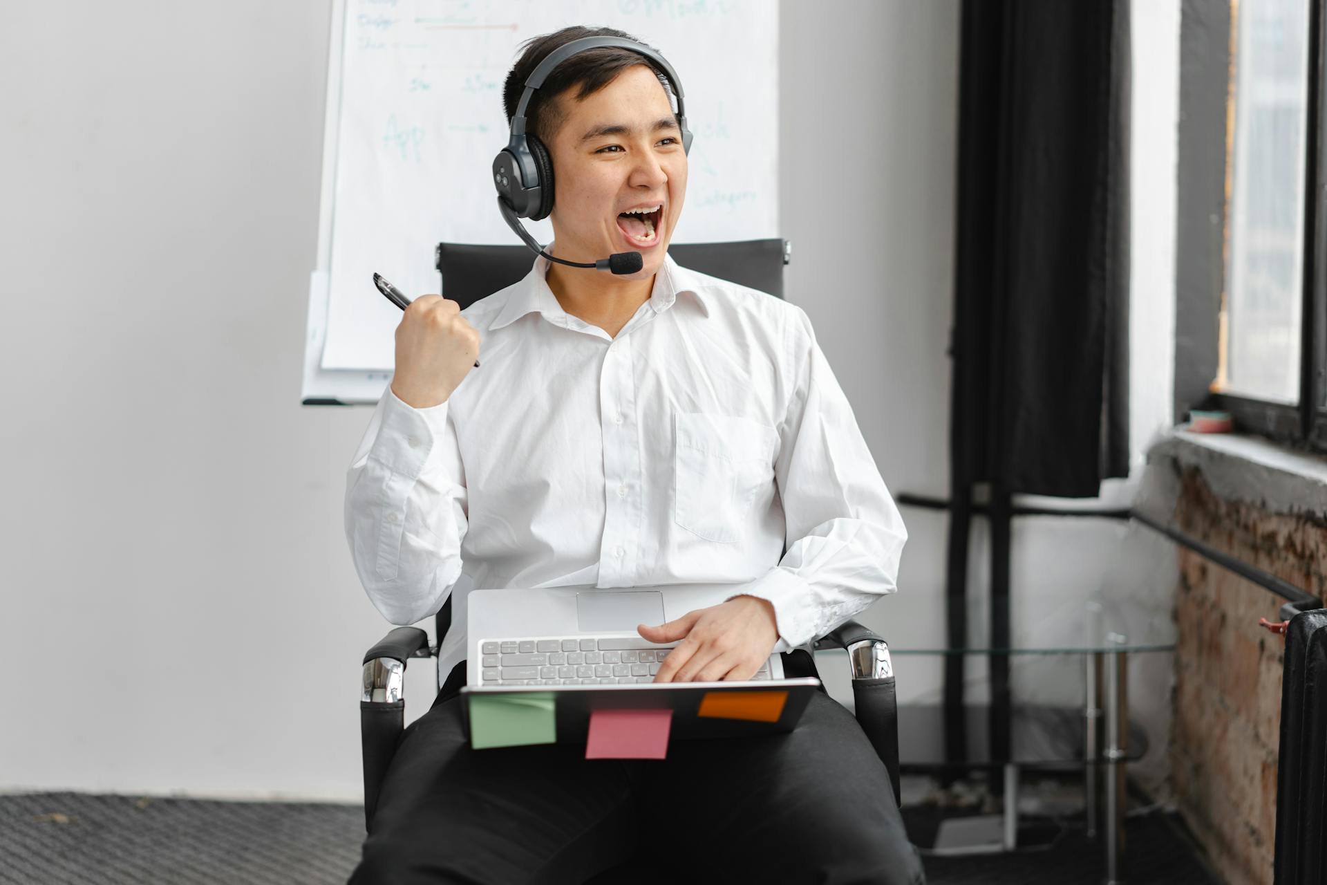 A happy young man with a headset is celebrating success during an online meeting in a modern office setting.