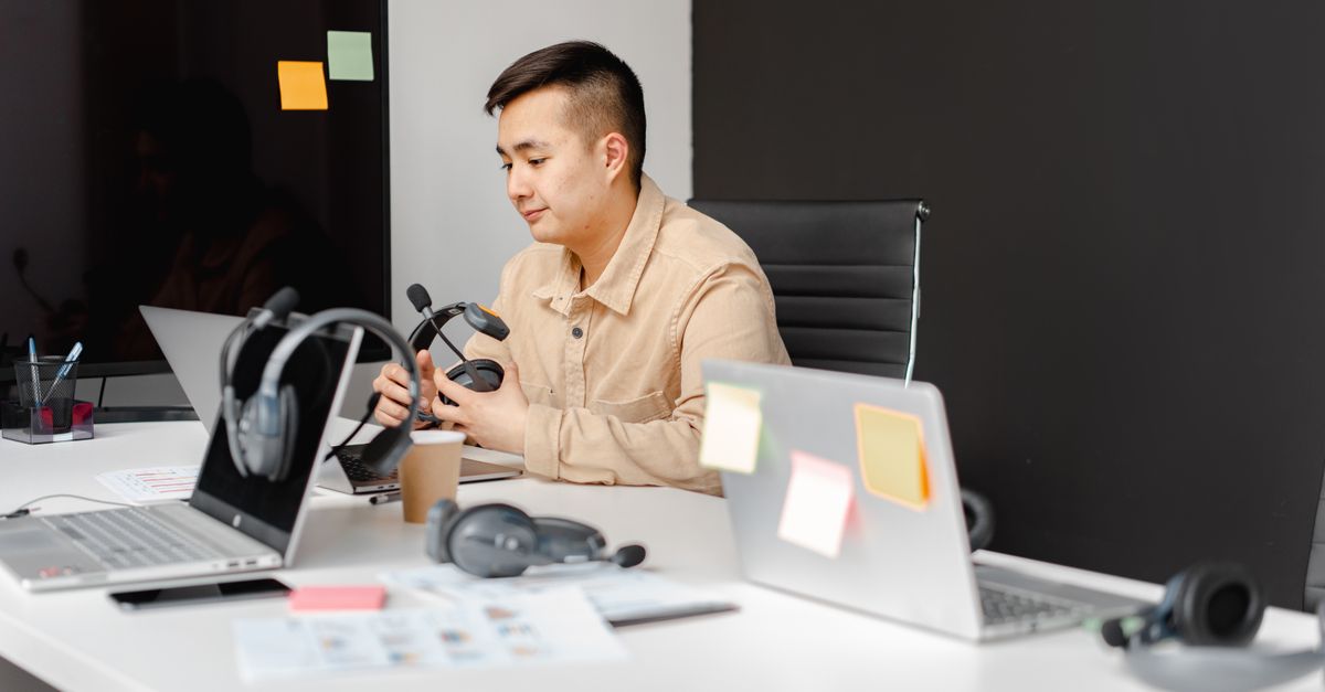 A person sitting at a cluttered desk with a laptop, sticky notes on the wall, and a cup of coffee, looking thoughtfully at the screen
