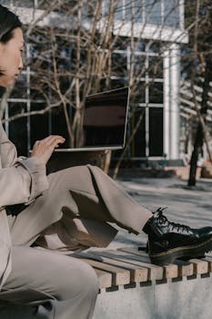 A woman in a trendy outfit works on her laptop while sitting outdoors.