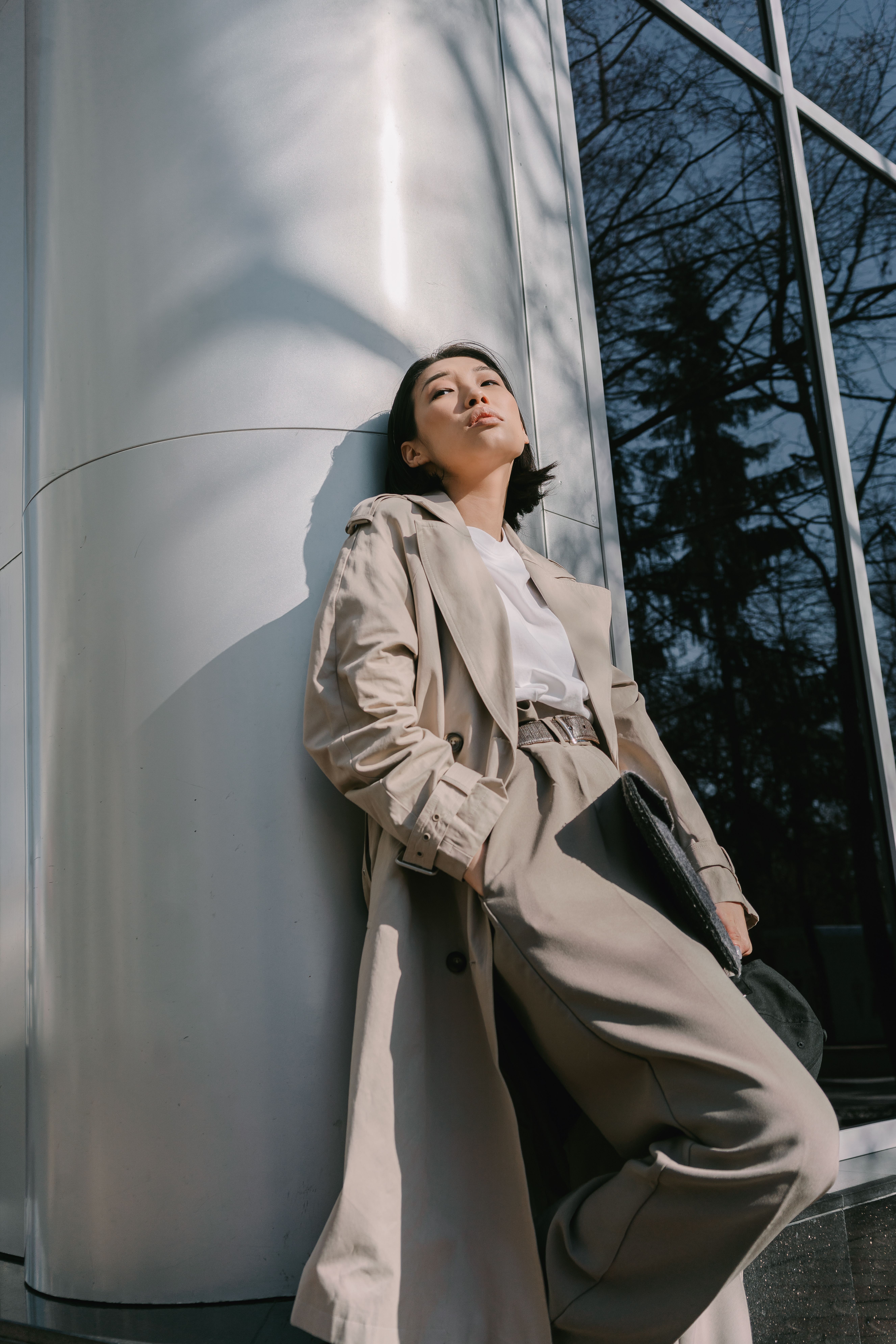 Chic Asian woman in a trench coat posing against a metal column in an urban setting.
