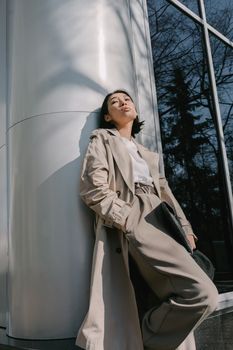 Chic Asian woman in a trench coat posing against a metal column in an urban setting.