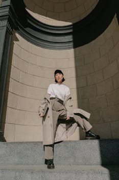 Fashionable woman in trench coat posing under sunlight on steps with stone wall background.