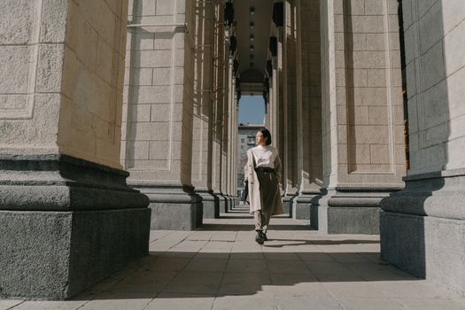 A fashionable woman in a trench coat walks through sunlit architectural columns.
