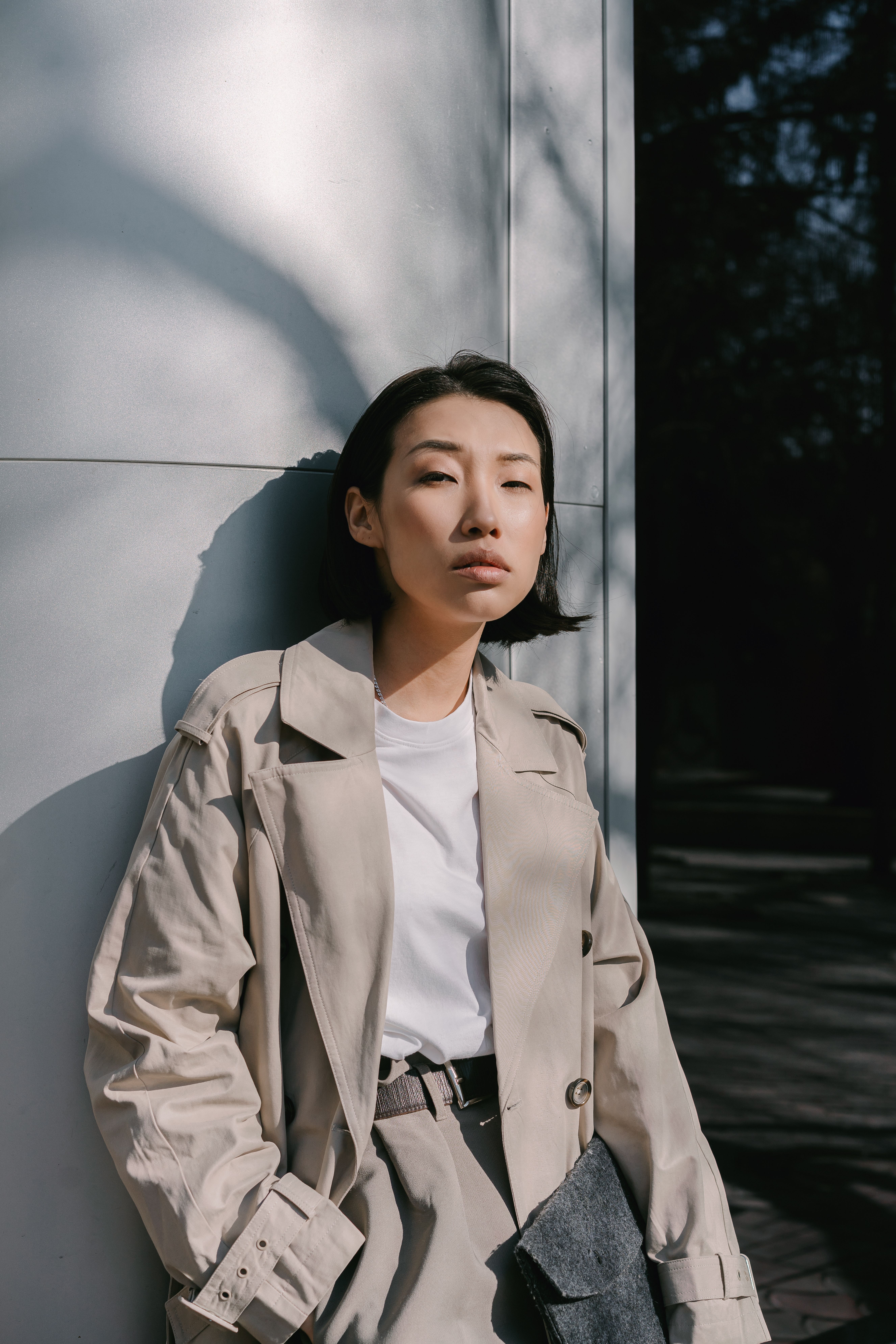 Fashionable Asian woman in a beige coat poses confidently against a minimalist wall in daylight.