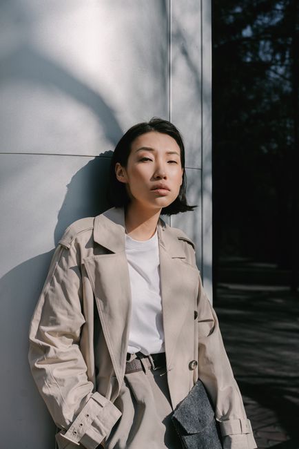 Fashionable Asian woman in a beige coat poses confidently against a minimalist wall in daylight.