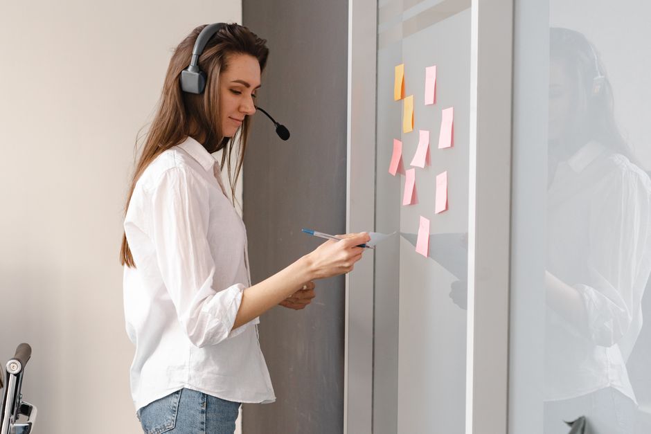 Woman wearing headset organizing tasks on a glass board with sticky notes in an office setting.