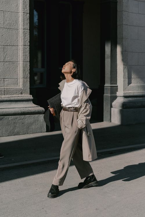 Free Fashionable woman in beige coat walking outdoors with eyes closed, basking in sunlight. Stock Photo