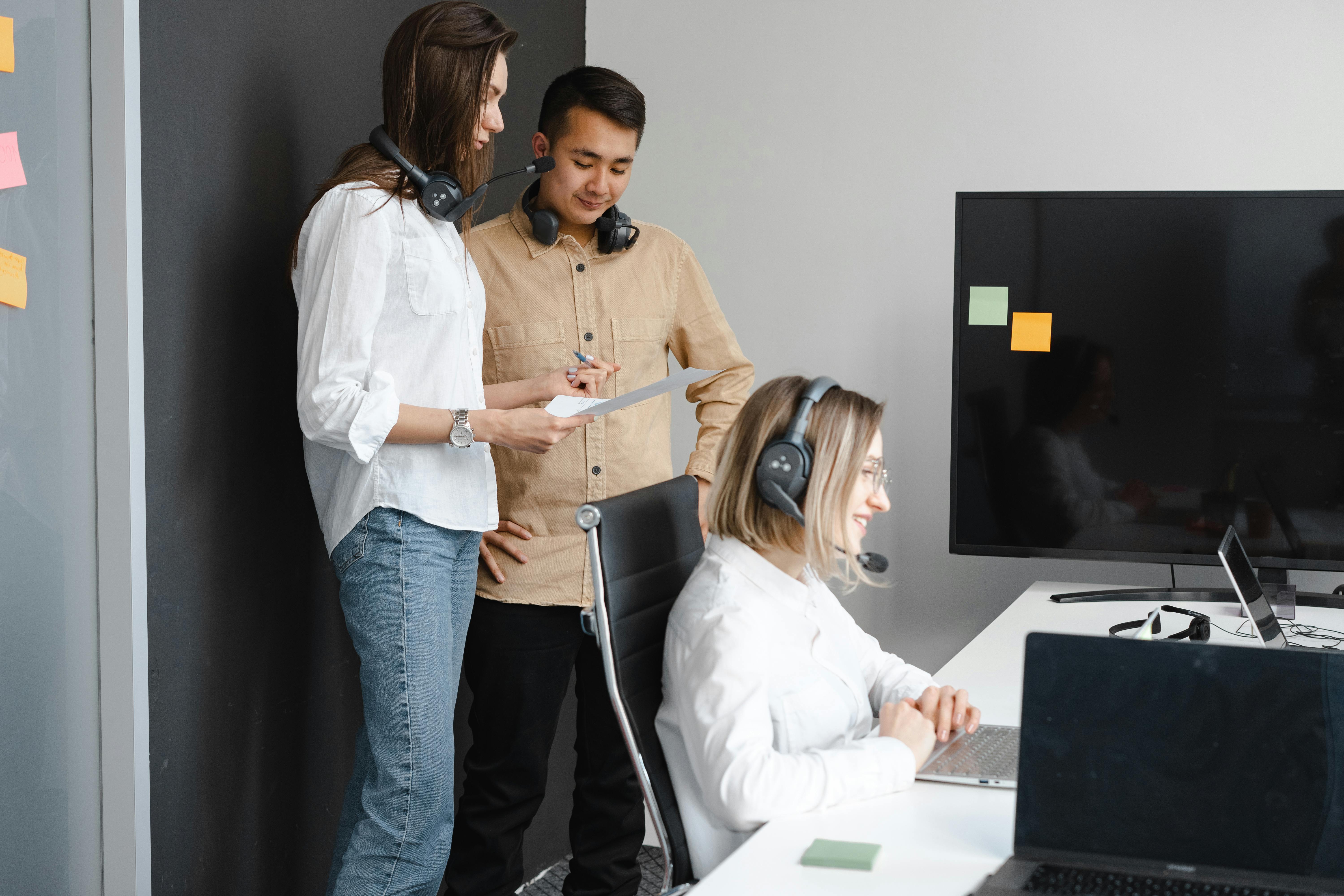 Three colleagues collaborate in an office, discussing documents and working on computers.