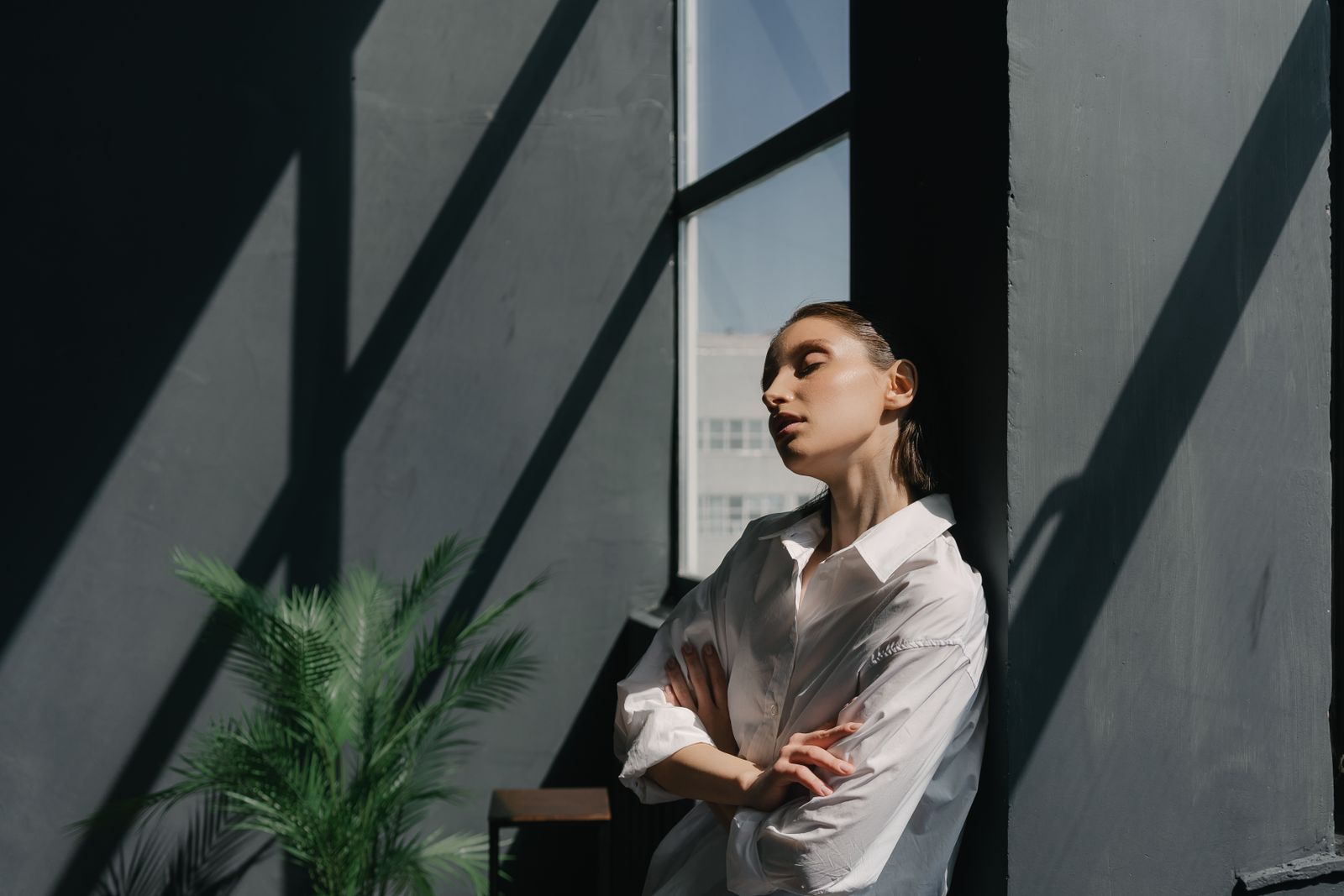Woman in white shirt leans against dark wall with eyes closed in angled sunlight beside a window