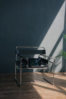 A modern metal and leather chair holding a tablet with a stock market graph against a dark wall. Natural light suggests an indoor setting.