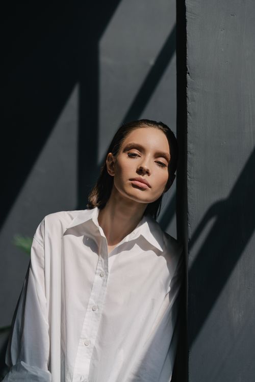 Free Confident woman in a white shirt leans against a black wall in soft daylight. Stock Photo