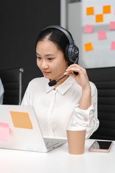 Asian woman using a laptop and headset for customer support in an office environment.