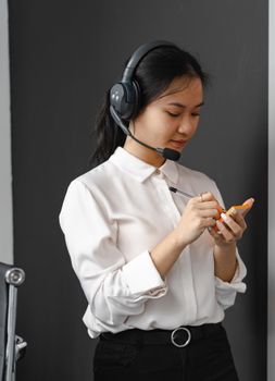 Asian woman in a white shirt with a headset writing notes, representing customer service.