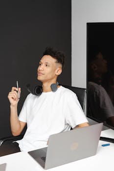 A young adult man seated indoors at a desk with a laptop and headset, engaging in thought.