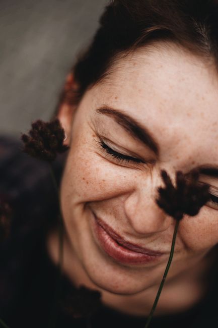 Positive young female with freckles and closed eyes near dark plants with fluffy inflorescence in daytime
