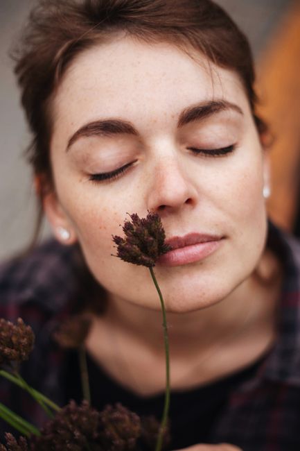 Thoughtful young lady with freckles and closed eyes with dark plants with fluffy inflorescence on face in daylight