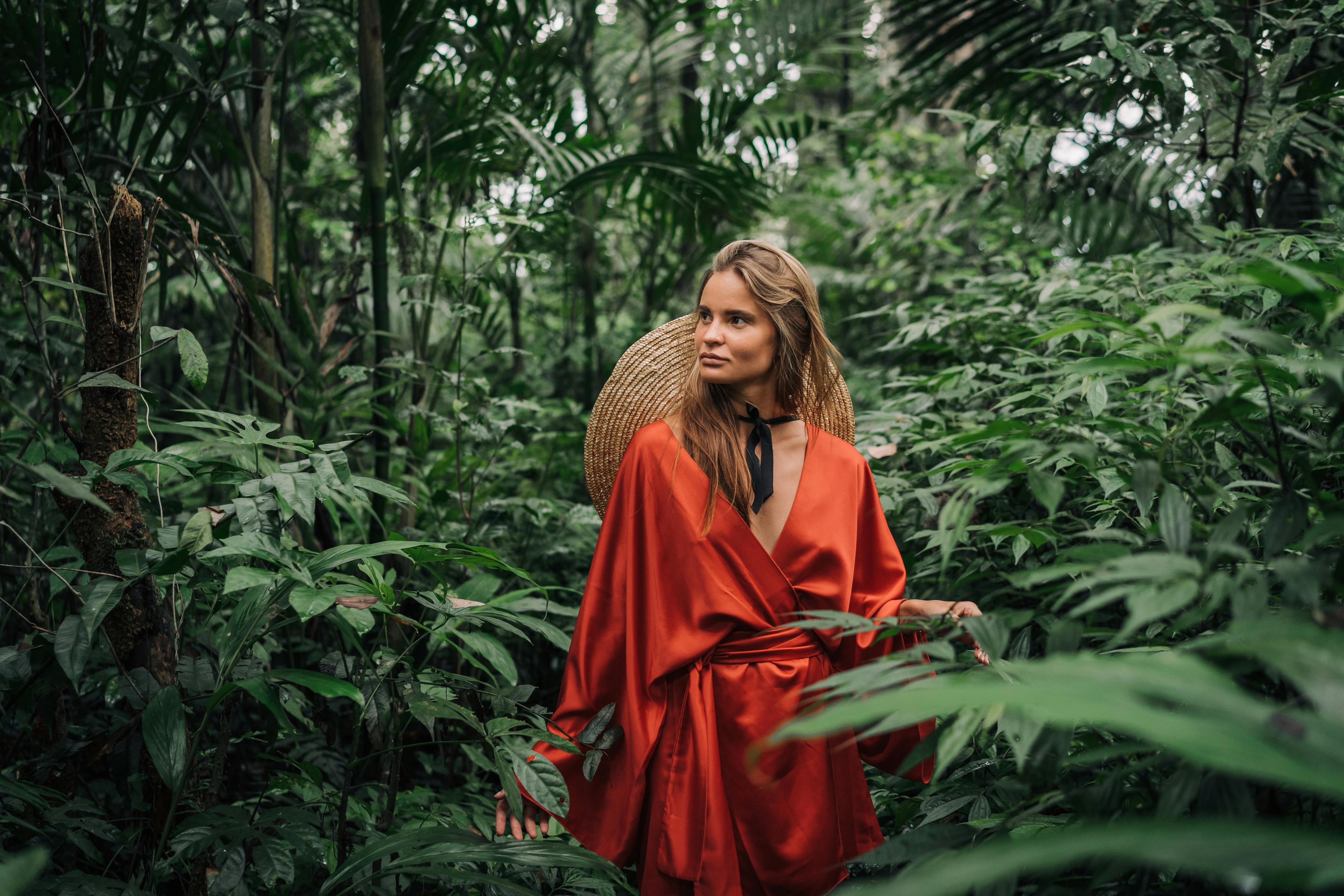 一个穿着红色长袍的时尚女人在郁郁葱葱的丛林绿地中摆姿势，创造了一个充满活力和异国情调的场景