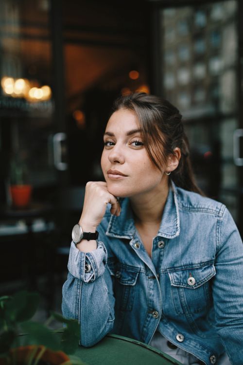 Free Dreamy young lady in casual clothes sitting at table in street cafeteria near glass doors while looking at camera in daytime Stock Photo