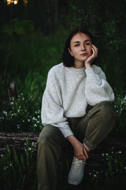 Woman in knit sweater sitting thoughtfully outdoors on a grass-covered tree trunk.
