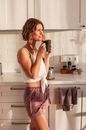 Side view of smiling lady in sleepwear standing in light kitchen while drinking cup of fresh beverage in morning near counter and cabinets
