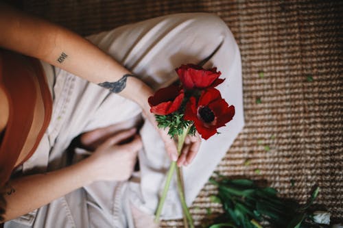 From above of crop anonymous person in casual clothes sitting with bouquet of red poppy flowers on carpet