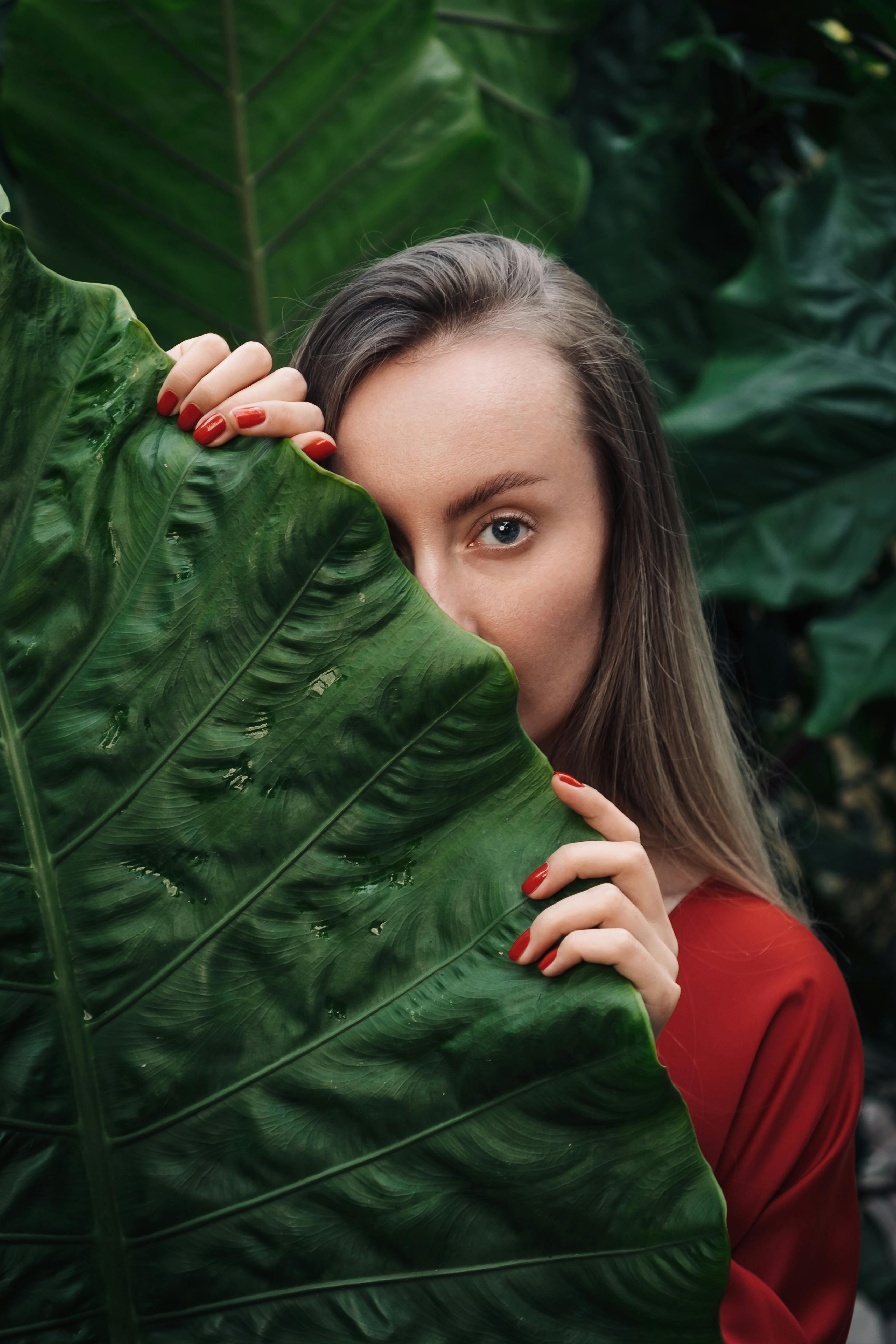 A woman in a red top peeks from behind a large tropical leaf, set in a lush green outdoor environment.
