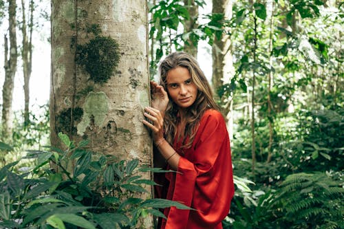 Woman in Red Long Sleeve Shirt Leaning on Brown Tree Trunk