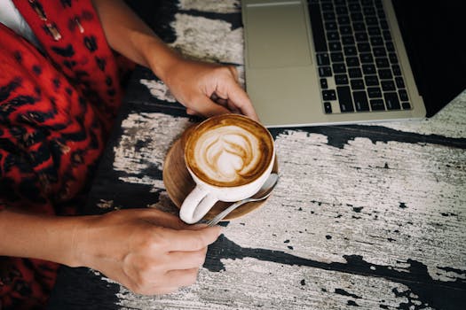 A person holding a coffee with latte art beside a laptop on a rustic table. Cozy scene.