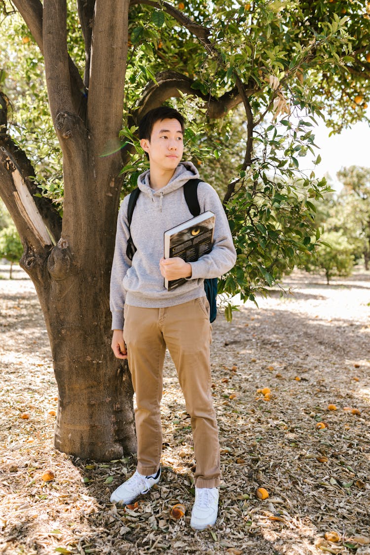 College Boy Student Standing Near The Tree While Holding A Book