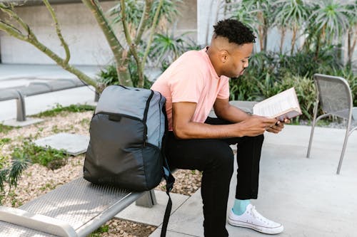 Free Man in Pink Crew Neck T-shirt and Black Pants Sitting on Black Chair Reading Book Stock Photo