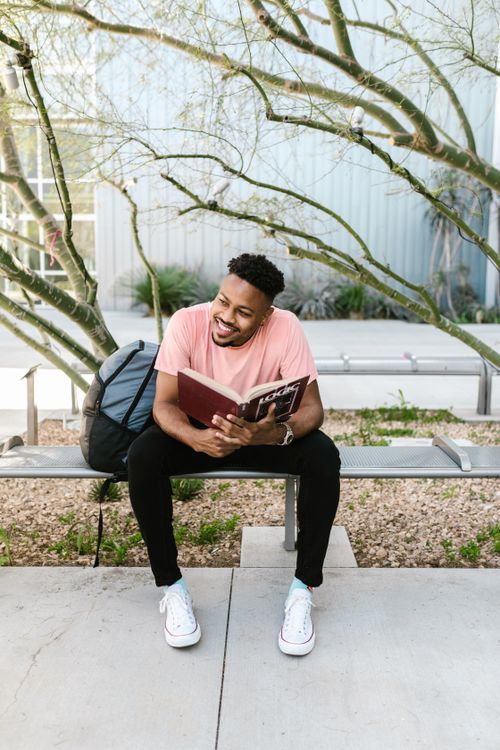 Free Casual young man reading outdoors on campus bench with backpack. Stock Photo