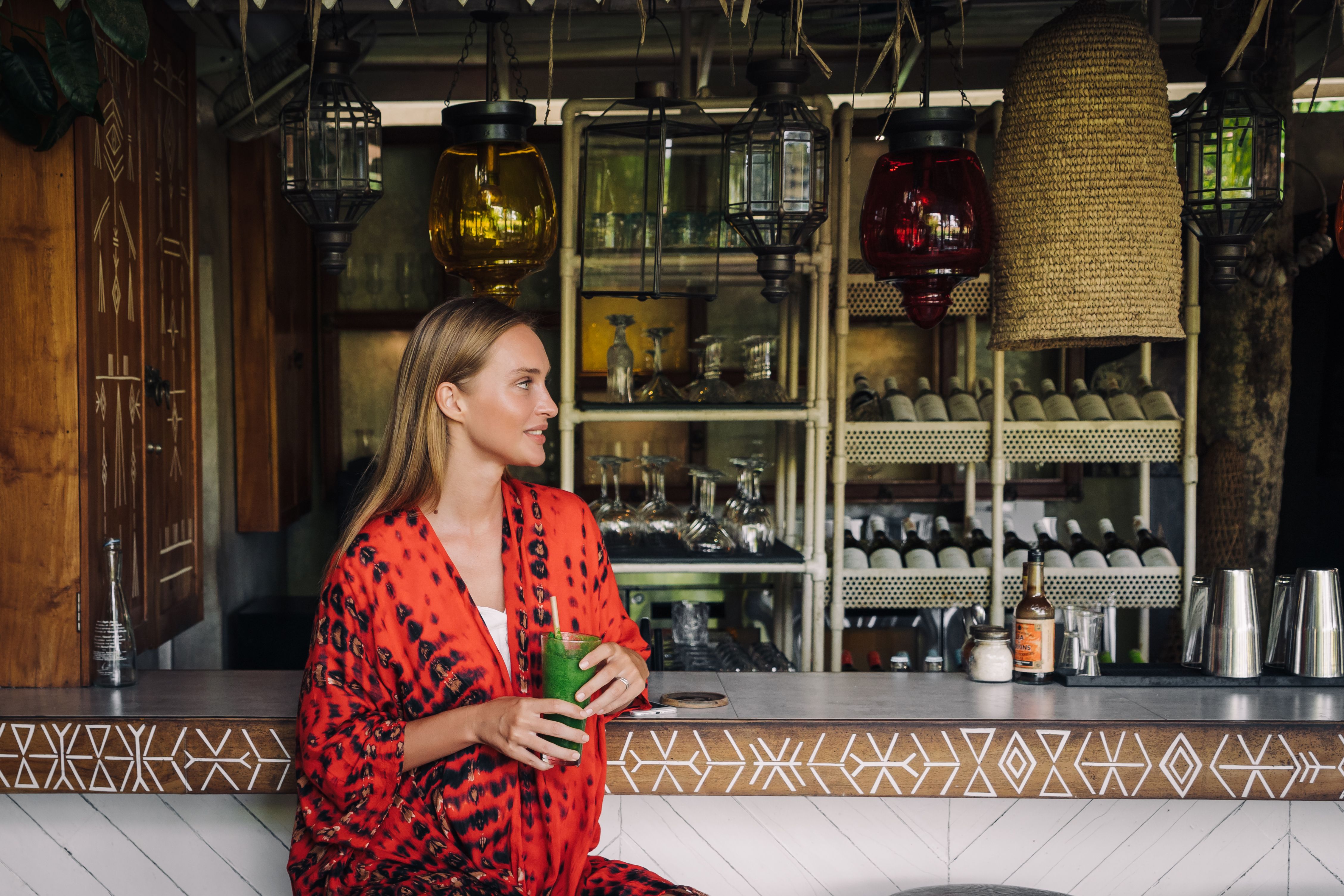 Woman in red patterned robe having a green smoothie at an elegant indoor bar.