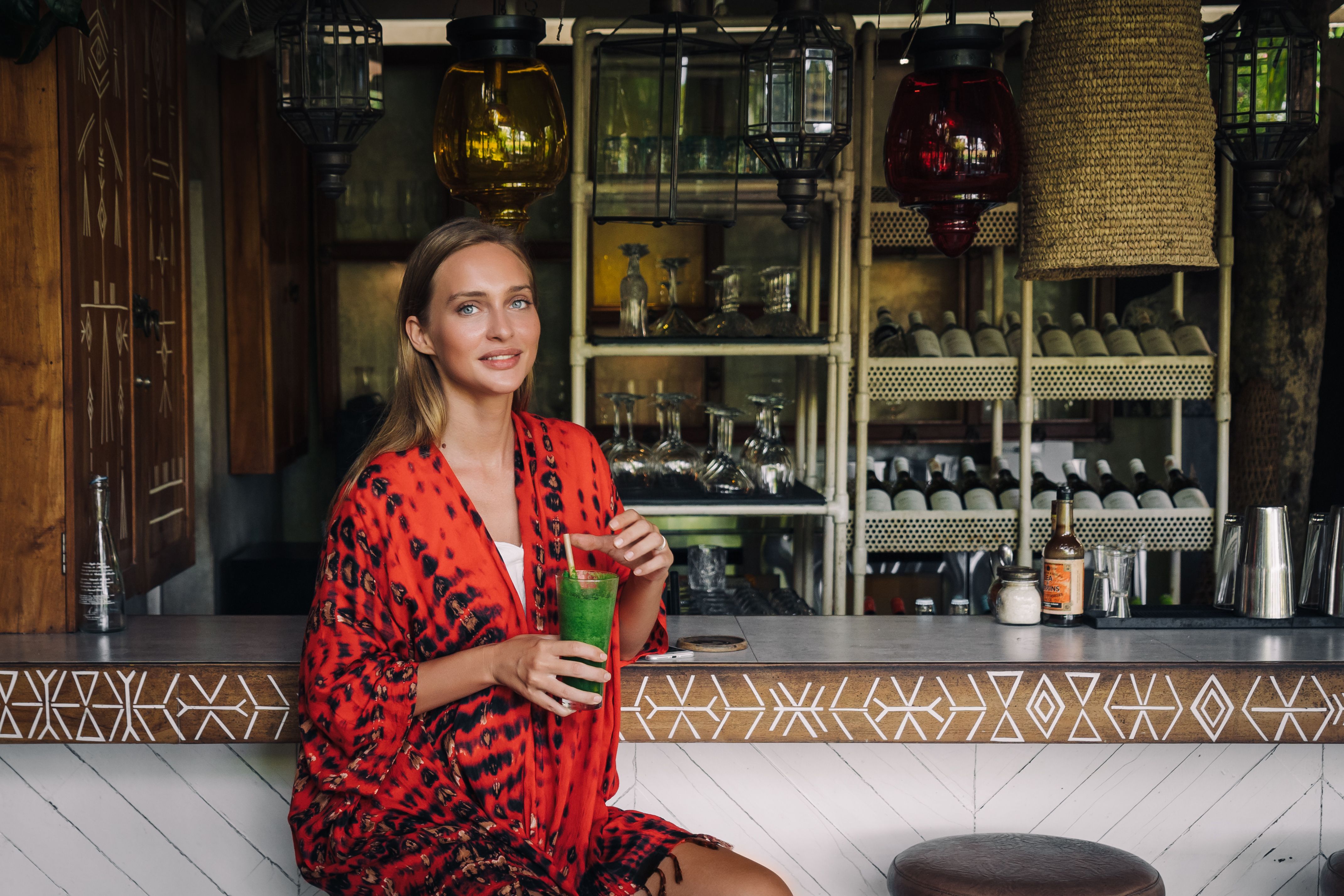 Woman in a vibrant robe holding a green smoothie at a trendy bar with elegant decor.