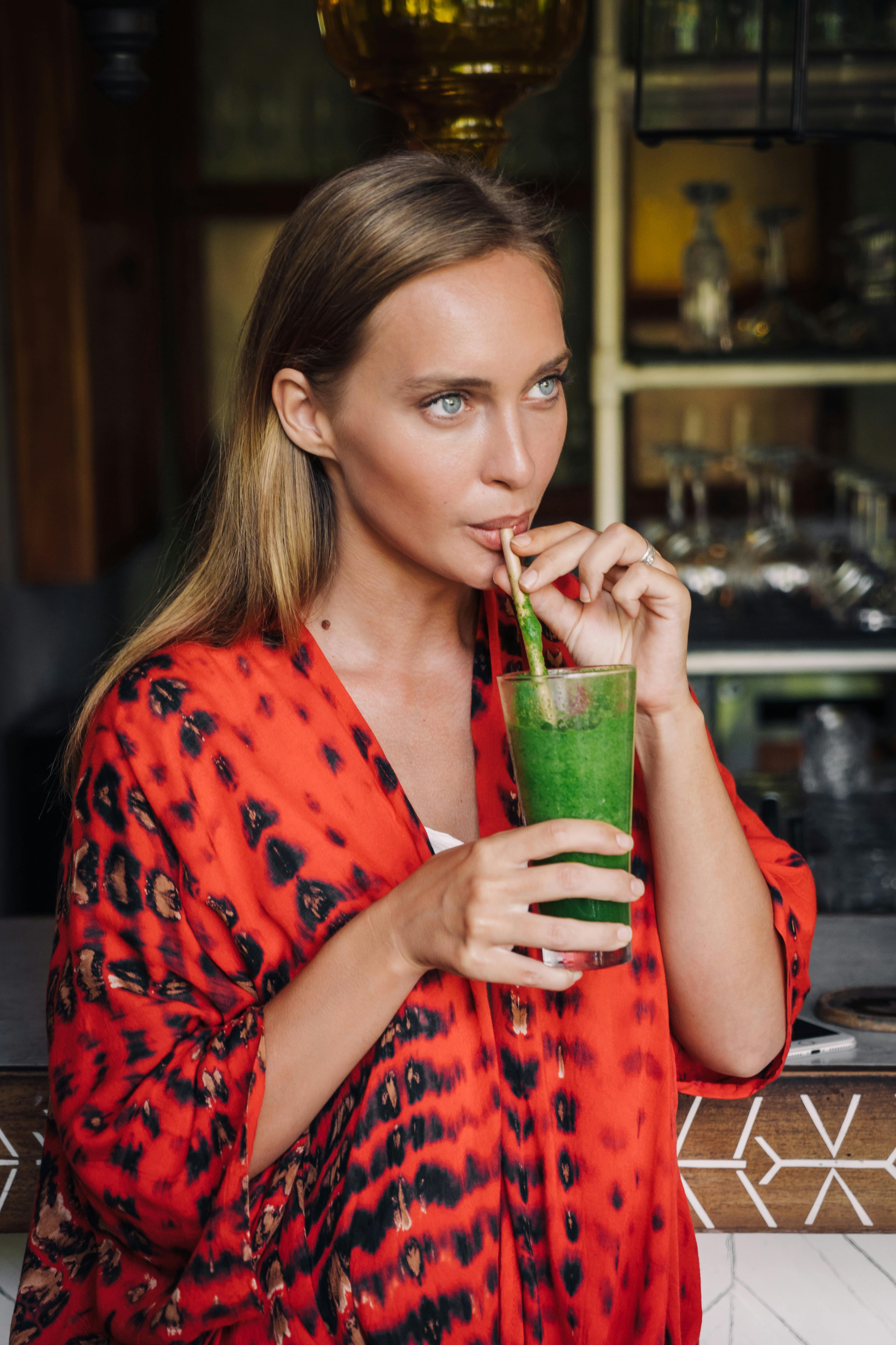 Stylish woman in vibrant red attire sipping on a fresh green smoothie at a modern indoor cafe.