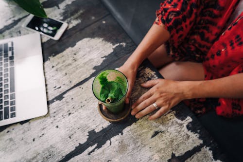 A Person Holding a Green Smoothie Beside the Laptop