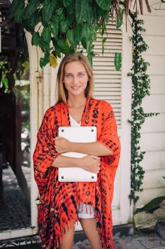 Smiling woman in orange shawl holding a laptop outside a charming home.