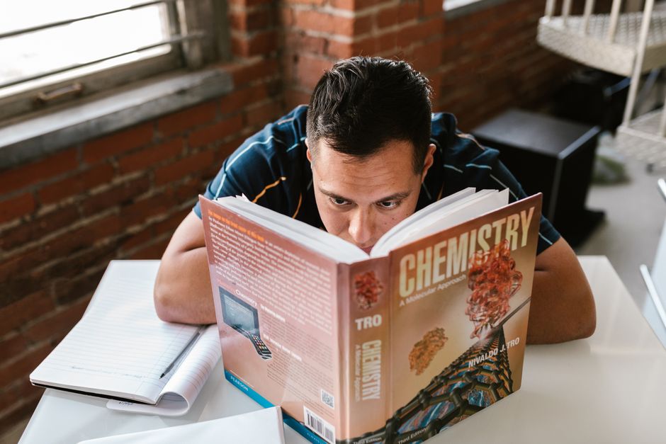 Adult male studying a chemistry textbook at a desk in a cozy, brick-walled study area. Adult male studying a chemistry textbook at a desk in a cozy, brick-walled study area.