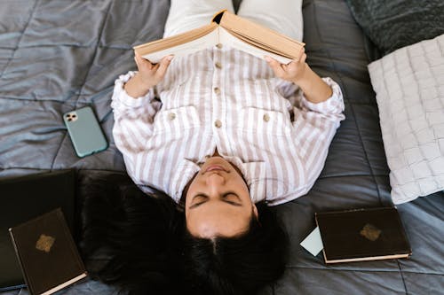 A Young Woman Reading a Book while Lying Down on a Bed