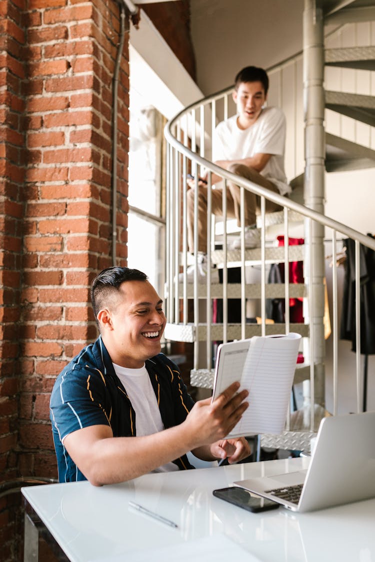 A Happy Young Man Holding A Notebook While Sitting At A Desk