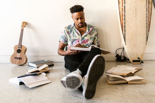 Free A young man reads a book while sitting among books and a ukulele, with a surfboard leaning on the wall. Stock Photo