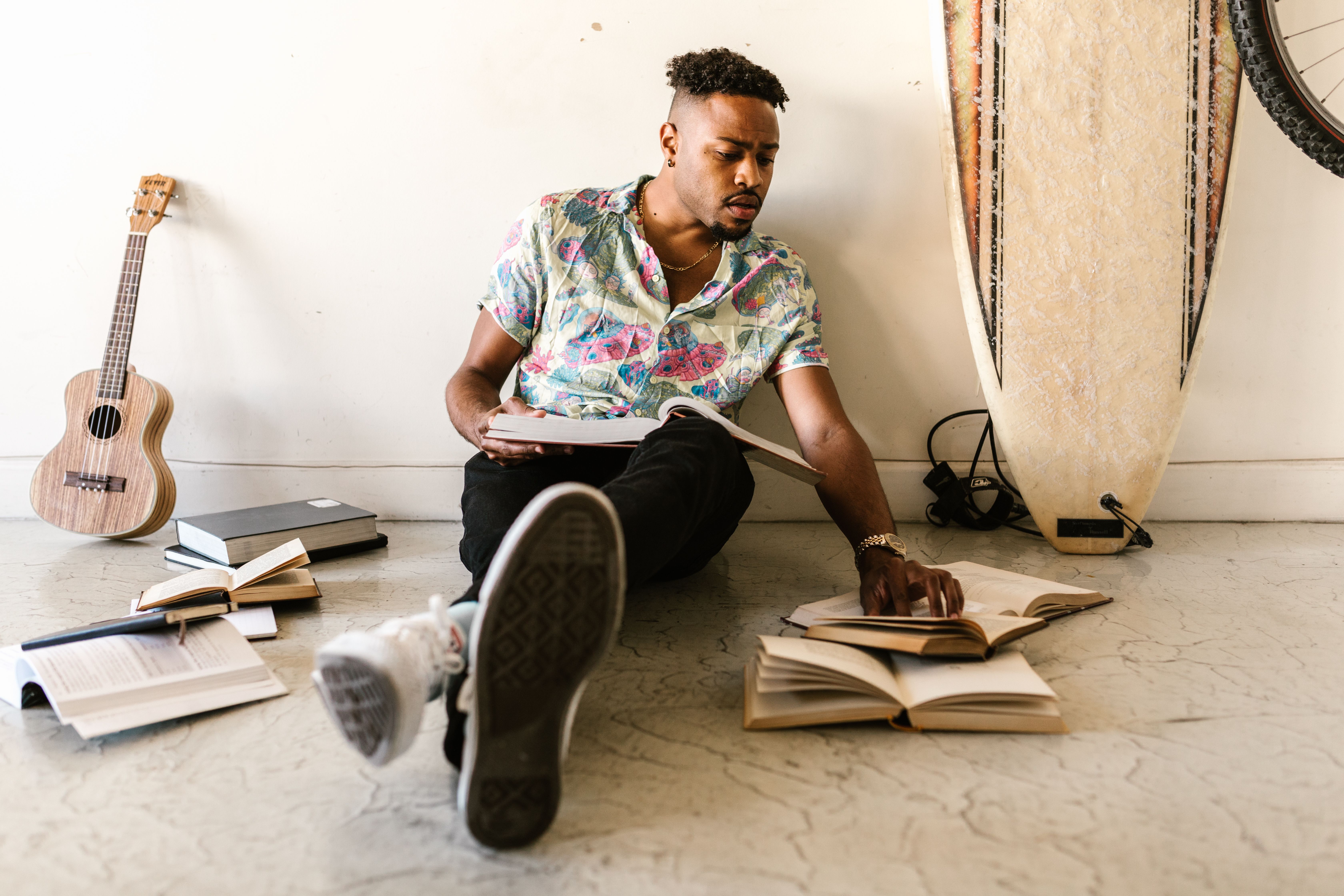 A young man sitting on the floor reading books, surrounded by a guitar and surfboard, embracing a creative and scholarly