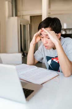 Anxious teenager studying at a desk with a laptop and open book, reflecting stress and focus.
