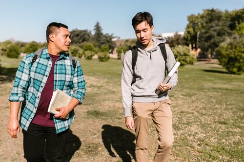Free Two male college students walk on a sunny campus day holding textbooks and backpacks. Stock Photo
