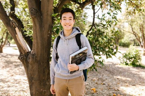 Free Young college student standing outside holding books, smiling at the camera. Stock Photo